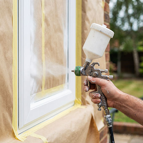 A close up image of a male hand holding a HVLP spray gun spraying paint onto a UPVC window frame outdoors.