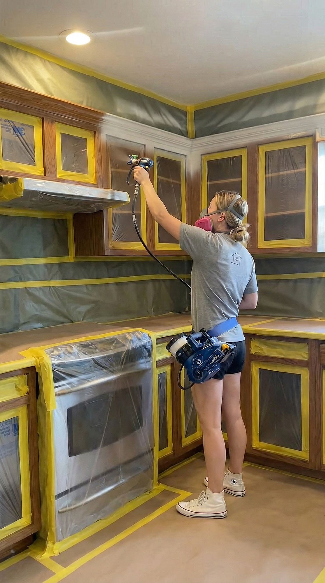 A kitchen spray painter spraying primer onto brown wooden cabinets that have been masked off.
