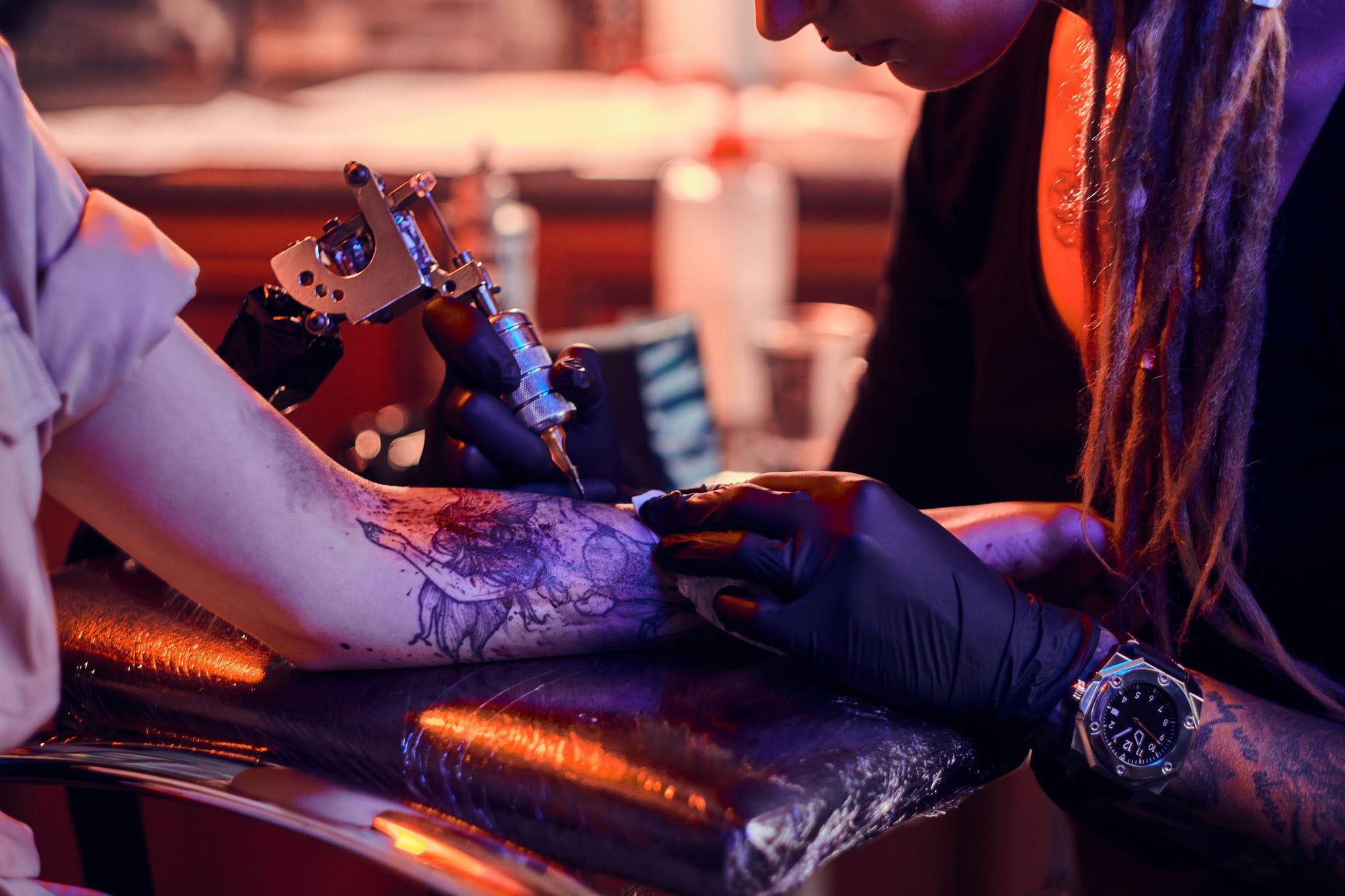 Tattoo artist wearing black gloves tattooing a client's forearm